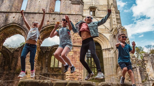Four children jump for joy from a wall of the ruin at Fountains Abbey